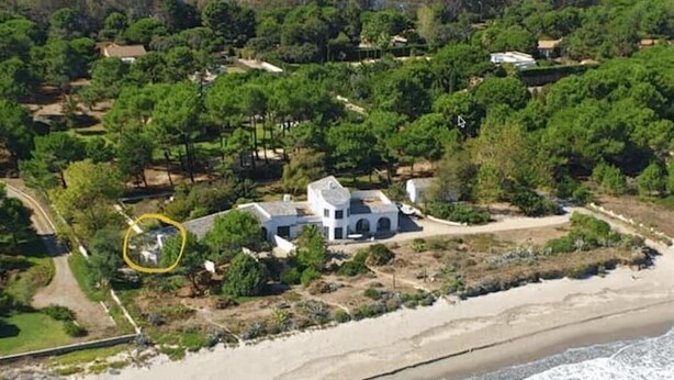 Vue aérienne d’une propriété située sur la côte, avec un bâtiment blanc entouré de verdure et une plage.