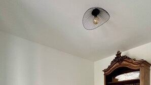 Una cama bien hecha con sábanas blancas y toallas enrolladas, un armario de madera con ropa colgada, una lámpara en una mesita de noche y una lámpara de techo original.