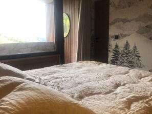 Un lit recouvert d’une couette blanche à texture, un mur en pierre, une fenêtre donnant sur un paysage verdoyant et un mur orné d’une fresque représentant une forêt.