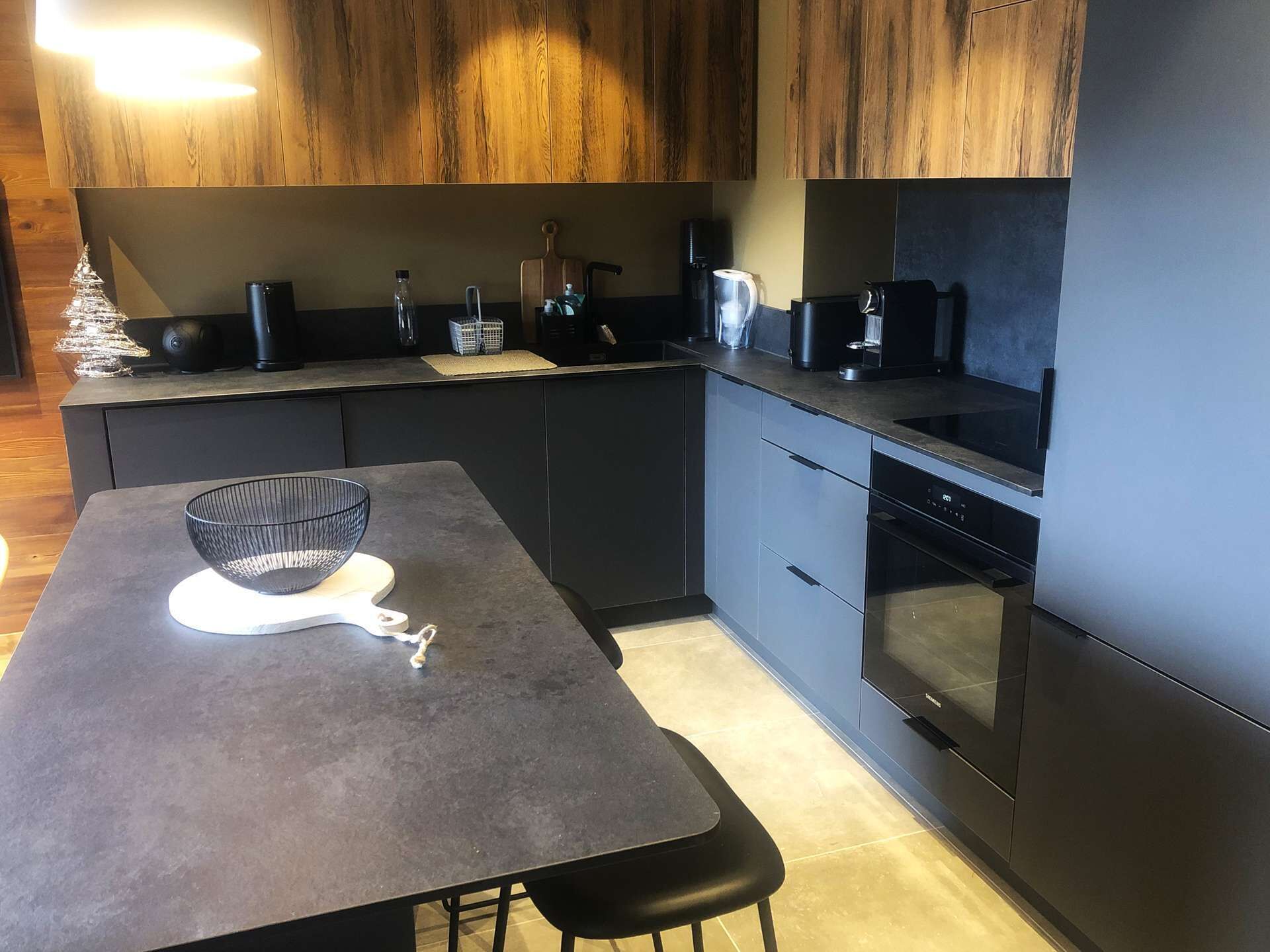 A modern kitchen with dark cabinetry, a built-in oven, and a dining area with a table and chairs.