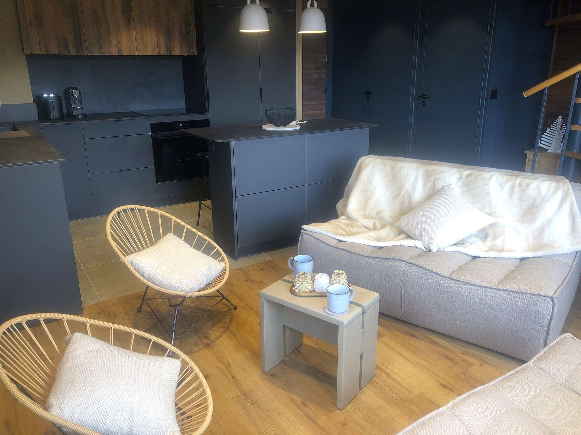 A modern living space with a grey sofa, two wicker chairs, a small side table, and a kitchen area with dark cabinets and a coffee machine.