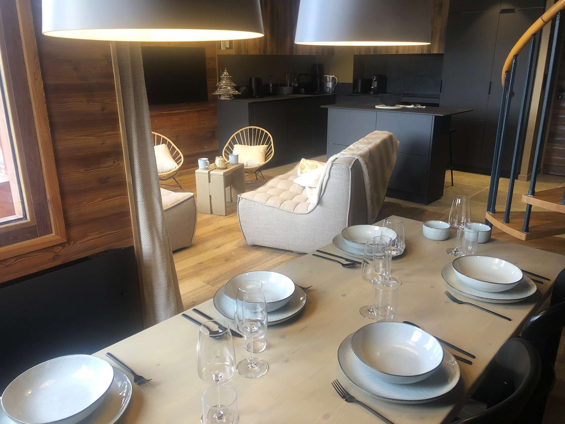 A dining area with a table set for six, including plates, glasses, and cutlery. There are two chairs with cushions, a small side table, and a lamp. The room has wooden flooring and a modern kitchen in the background.