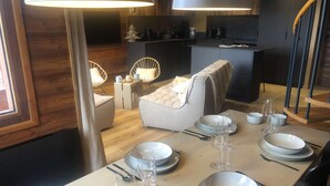 A dining area with a table set for six, including plates, glasses, and cutlery. There are two chairs with cushions, a small side table, and a lamp. The room has wooden flooring and a modern kitchen in the background. - Le Portail 13 - Outstanding apartment at the foot of the slopes (LES AVANCHERS VALMOREL)