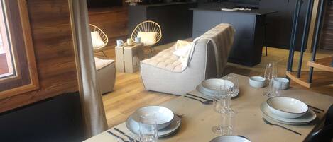 A dining area with a table set for six, including plates, glasses, and cutlery. There are two chairs with cushions, a small side table, and a lamp. The room has wooden flooring and a modern kitchen in the background.