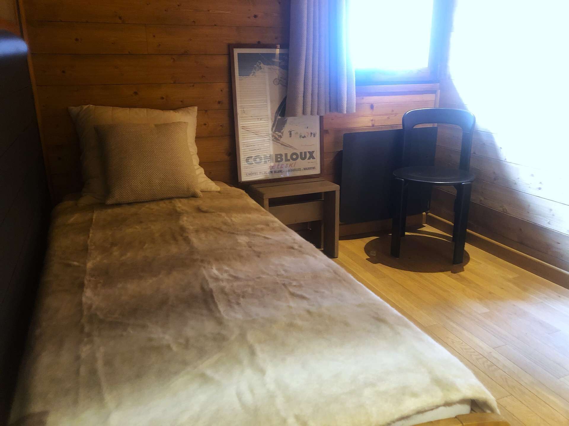 Un lit simple avec une couette blanche, une petite table de chevet en bois, une affiche encadrée et une chaise noire dans une pièce aux murs en bois et dotée d’une fenêtre avec des rideaux.