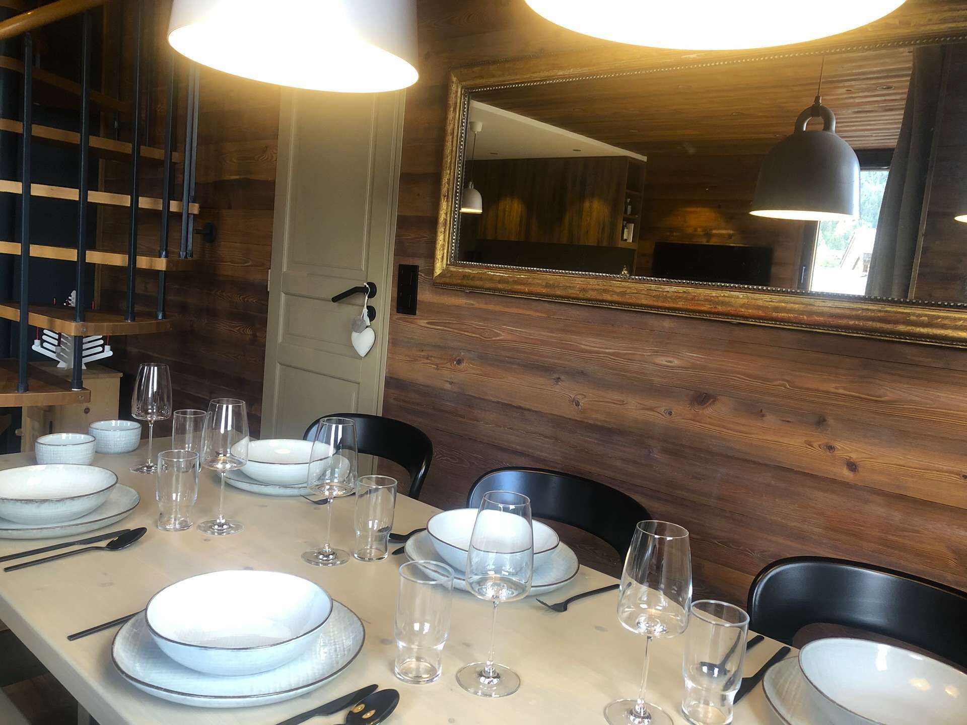 A dining area with a long table set for four, wooden walls, a mirror, and pendant lighting.