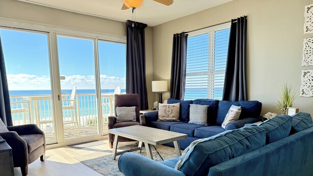 A living room with a blue sofa, a brown armchair, and a coffee table. There is a balcony with a view of the ocean.