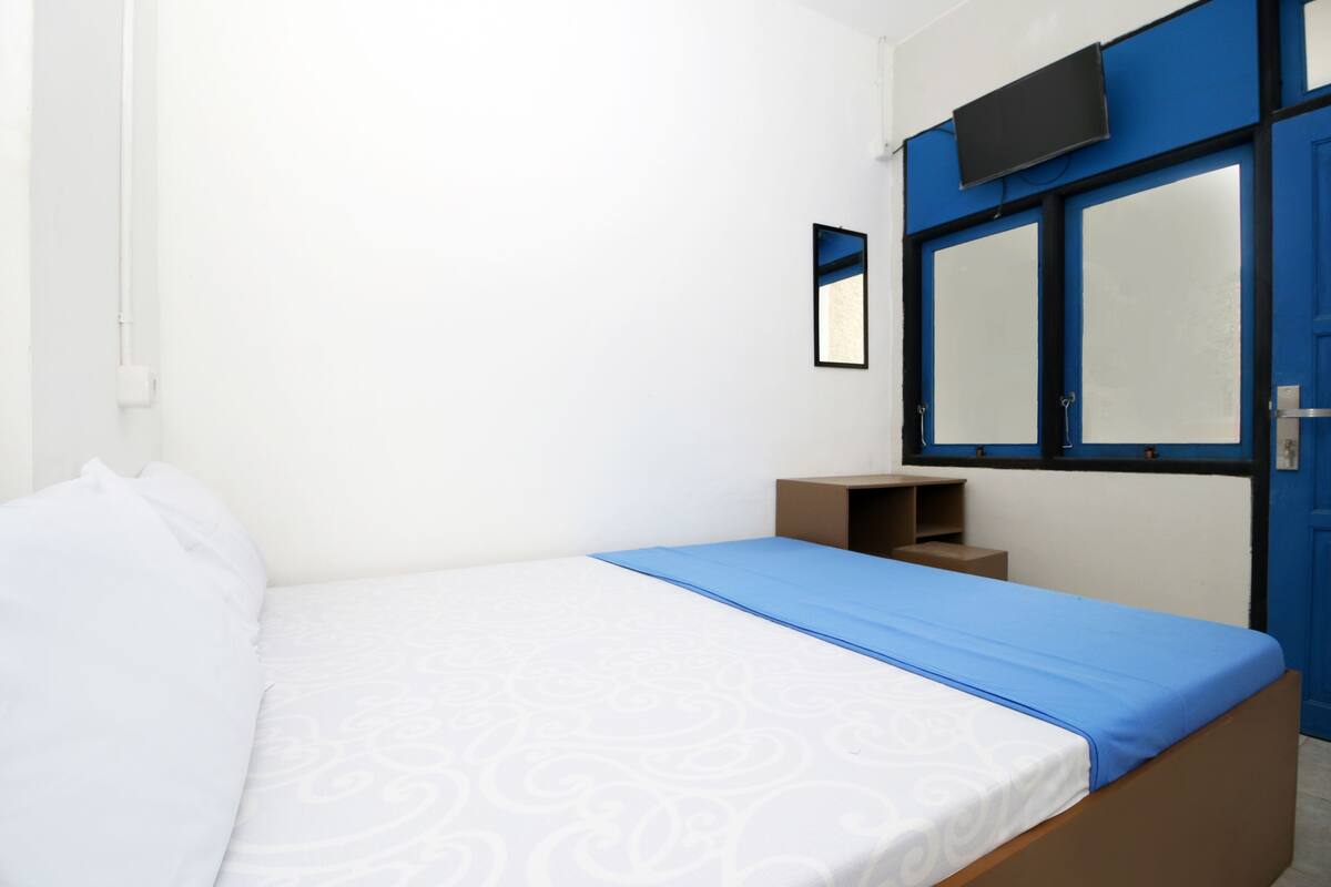 A bedroom with a bed, a blue bedspread, a white patterned bedspread, a small wooden nightstand, a mirror, and a blue door.