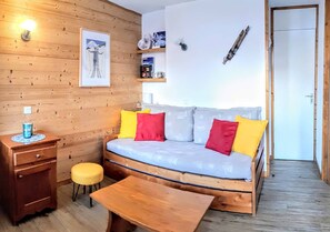 A cozy room with a wooden floor, a sofa with cushions, a small wooden table, a chair, a desk with a lamp, and a door.