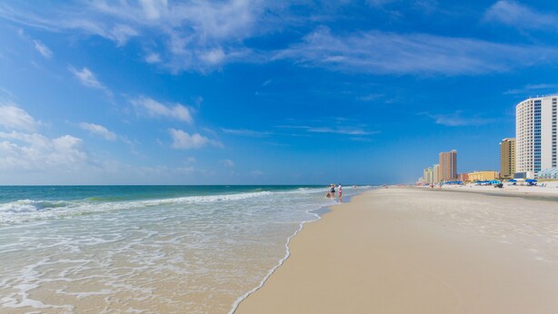Ein weiter Sandstrand mit sanften Wellen, blauem Himmel und hohen Gebäuden im Hintergrund.