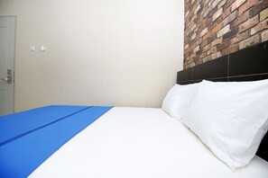 Una cama con sábanas blancas y una manta azul, una pared con diseño de ladrillo, y una puerta con pomo.