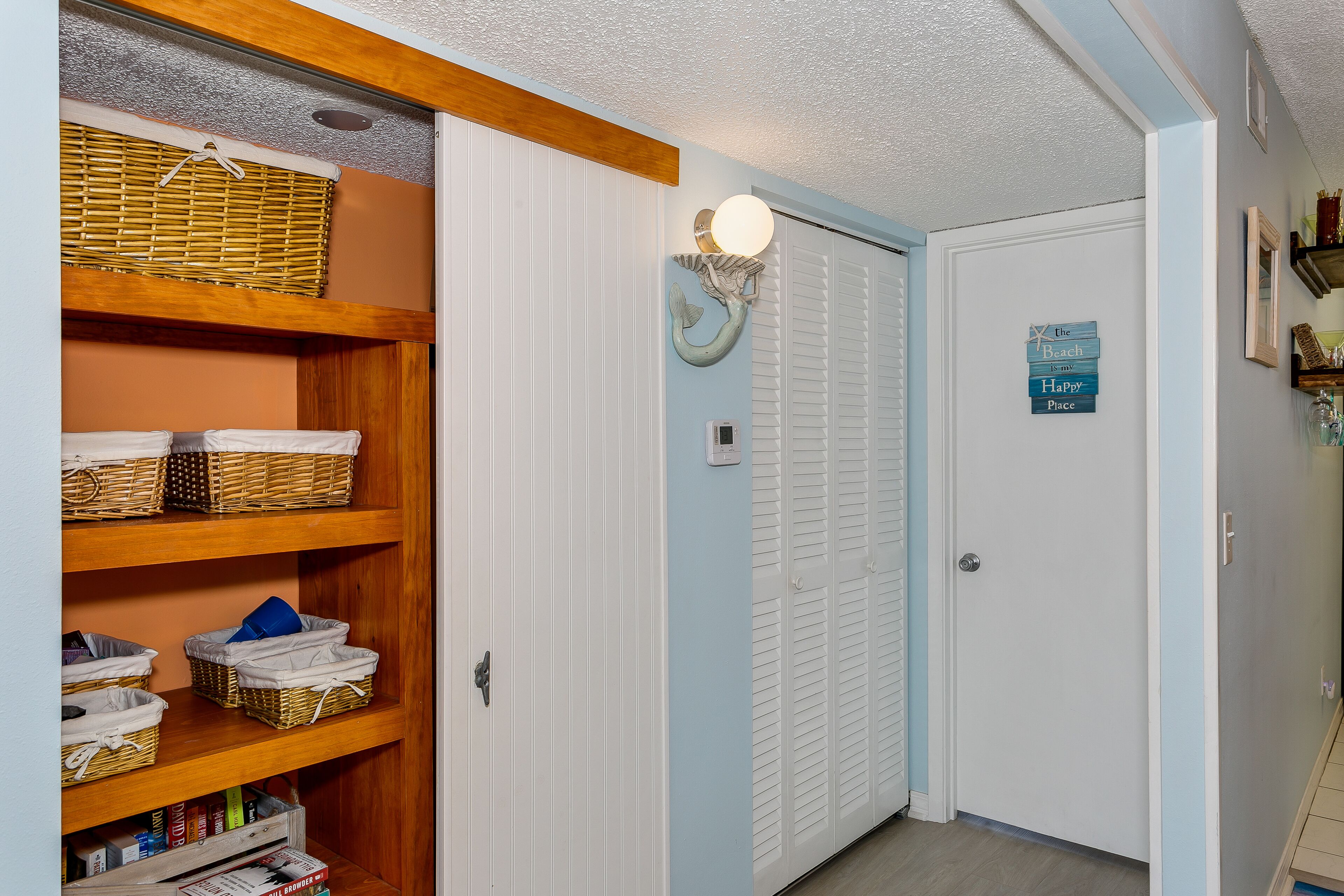 A hallway with a wooden shelf unit containing baskets and books, a white door with a light fixture above it, and a sign on the door.