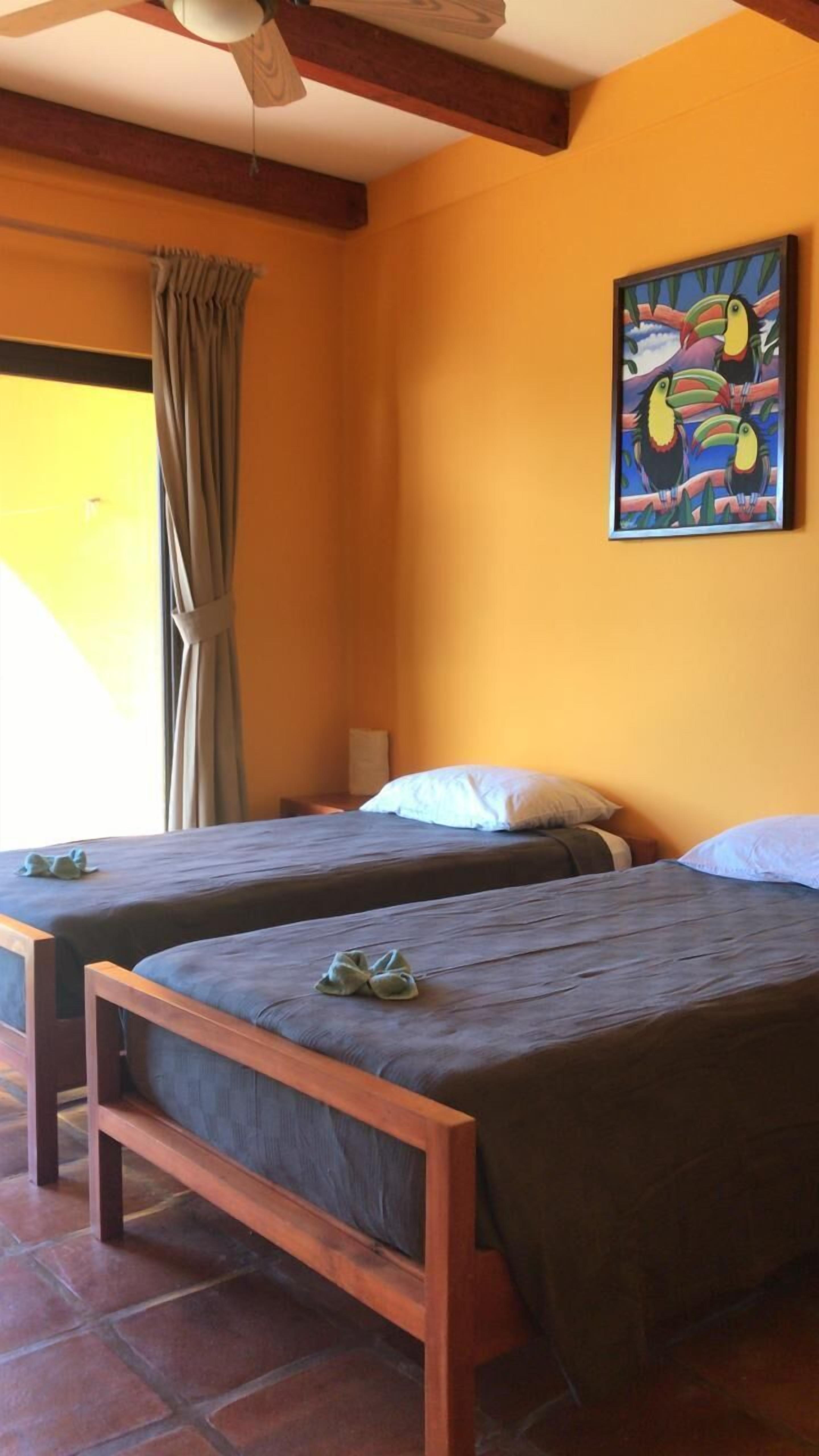 Una habitación con dos camas de madera, un cuadro de tucanes en la pared y una ventana con cortinas.