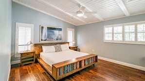Une chambre avec un lit en bois, un ventilateur de plafond et une bibliothèque placée sous le lit.