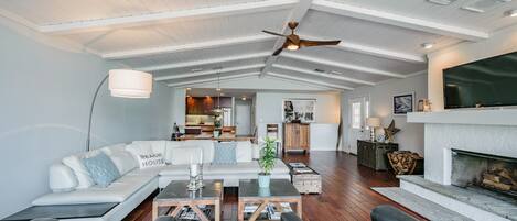 A spacious living room with a high ceiling, a fireplace, and a kitchen in the background.