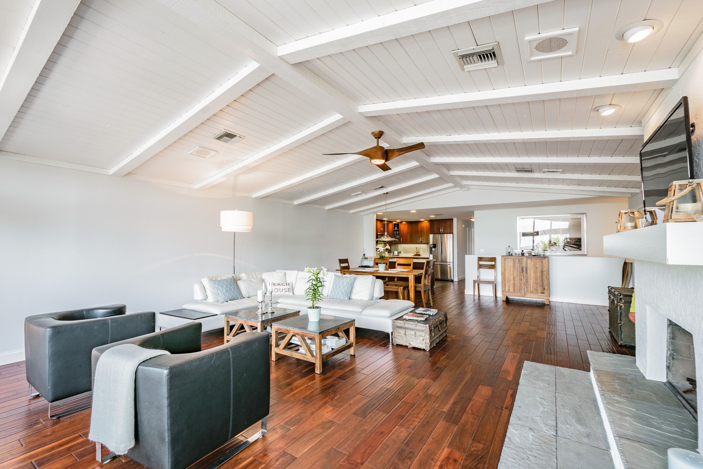 A spacious living room with a high ceiling, wooden flooring, a fireplace, and a kitchen in the background.