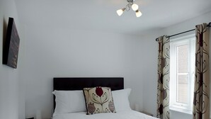 Een slaapkamer met een bed, hoofdeinde, nachtkastje, lamp en een raam met gordijnen.