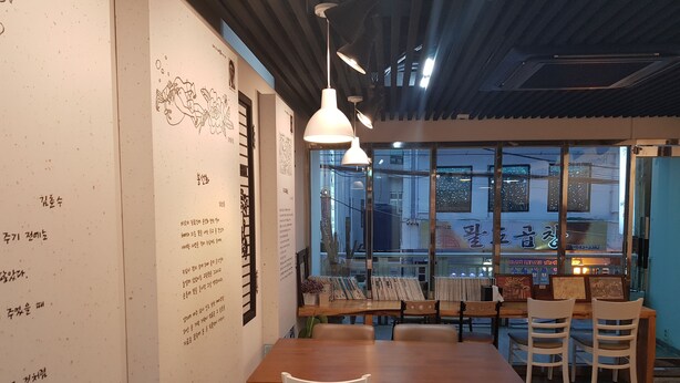 A dining area with a wooden table and white chairs, a wall with Korean text, and a view of the street through large windows. - Friends of Loft - Hostel (Tongyeong)
