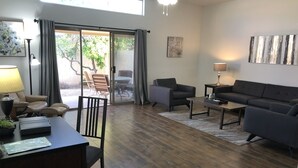 A modern living room with a sofa, chairs, a coffee table, and a desk. There is a sliding glass door leading to an outdoor area with a patio table and chairs. - One Story w/Htd Comm Pool & Spa: GOLF & Trails Nearby, Walk to Dine & Shop, Indoor Shuffleboard (Mesa)