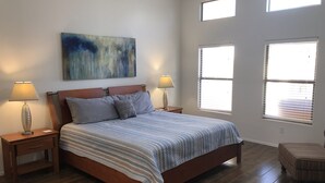 A bedroom with a wooden bed, two bedside tables with lamps, a large abstract painting on the wall, and three windows with blinds. - One Story w/Htd Comm Pool & Spa: GOLF & Trails Nearby, Walk to Dine & Shop, Indoor Shuffleboard (Mesa)