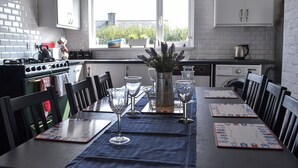 Un coin repas avec une table dressée pour quatre personnes, une fenêtre donnant sur un paysage verdoyant et une cuisine en arrière-plan.
