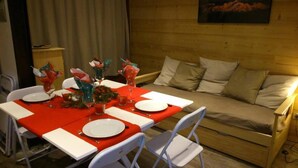 Un coin repas avec une table en bois dressée pour quatre personnes, des chaises blanches et un canapé en bois en arrière-plan.