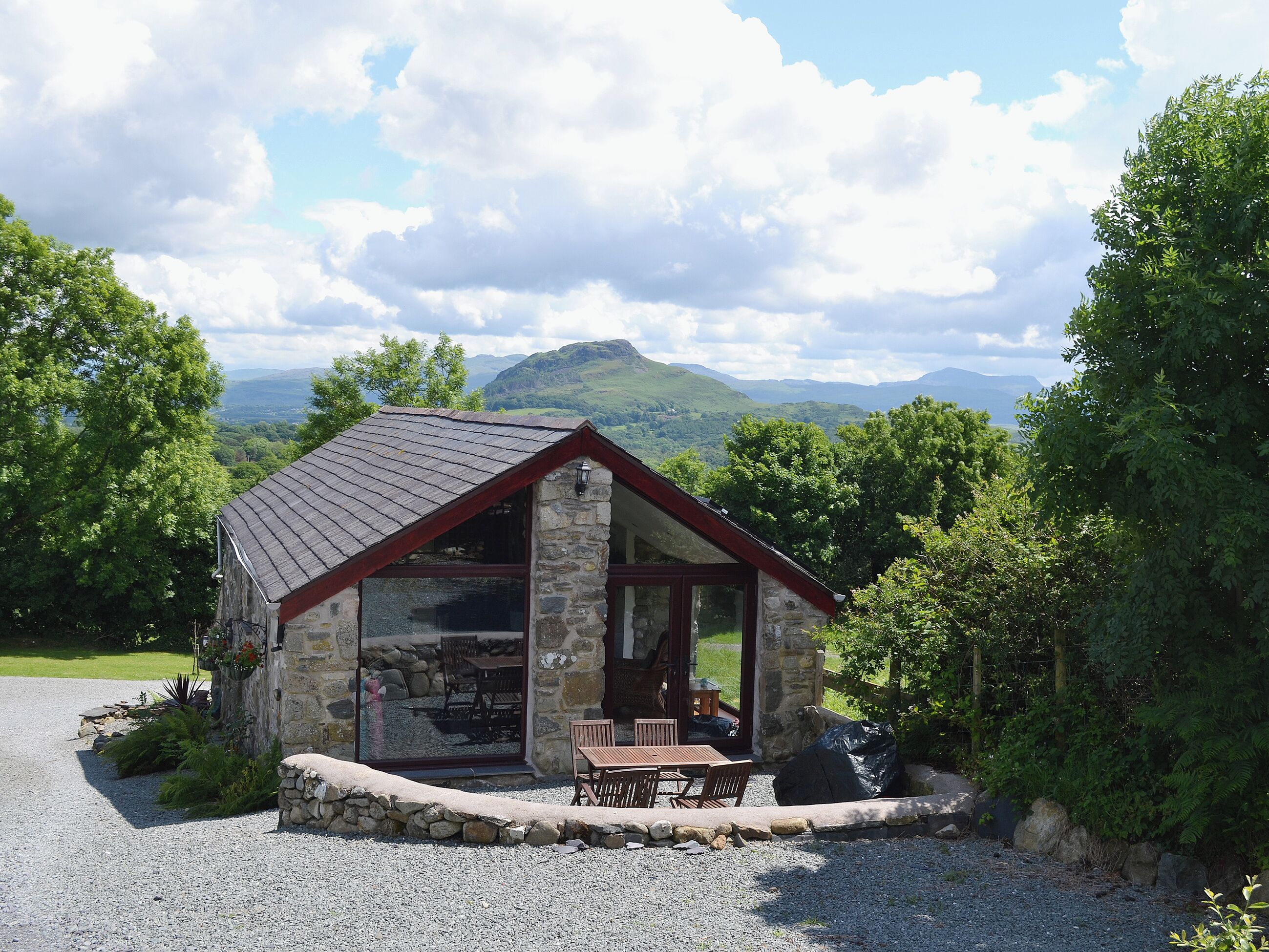 A stone cottage with a sloped roof, a glass door leading to a patio with a table and chairs, surrounded by a gravel driveway and lush greenery.