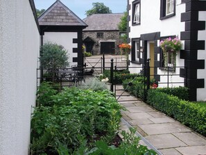A narrow pathway leading to a building with a black and white exterior, a gated entrance, and a small garden area with various plants.