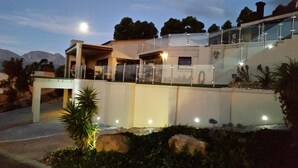 A modern house with a glass balcony, a stone wall, and a well-lit garden area. - Private Villa With Magnificent Sea Views  (Cape Town)