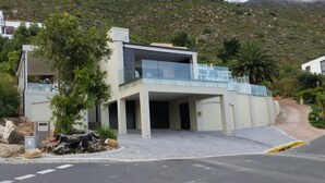 A modern two-story building with a glass balcony, a driveway, and a mountain backdrop. - Private Villa With Magnificent Sea Views  (Cape Town)
