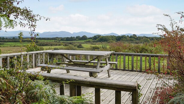 A wooden deck with picnic tables overlooking a scenic landscape with mountains in the distance. - 3 bedroom accommodation in Llanbedrgoch, near Benllech (Llanbedrgoch, near Benllech)