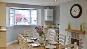 Een eetruimte met een houten tafel gedekt voor vier personen, witte stoelen, een vaas met bloemen en een klok aan de muur.