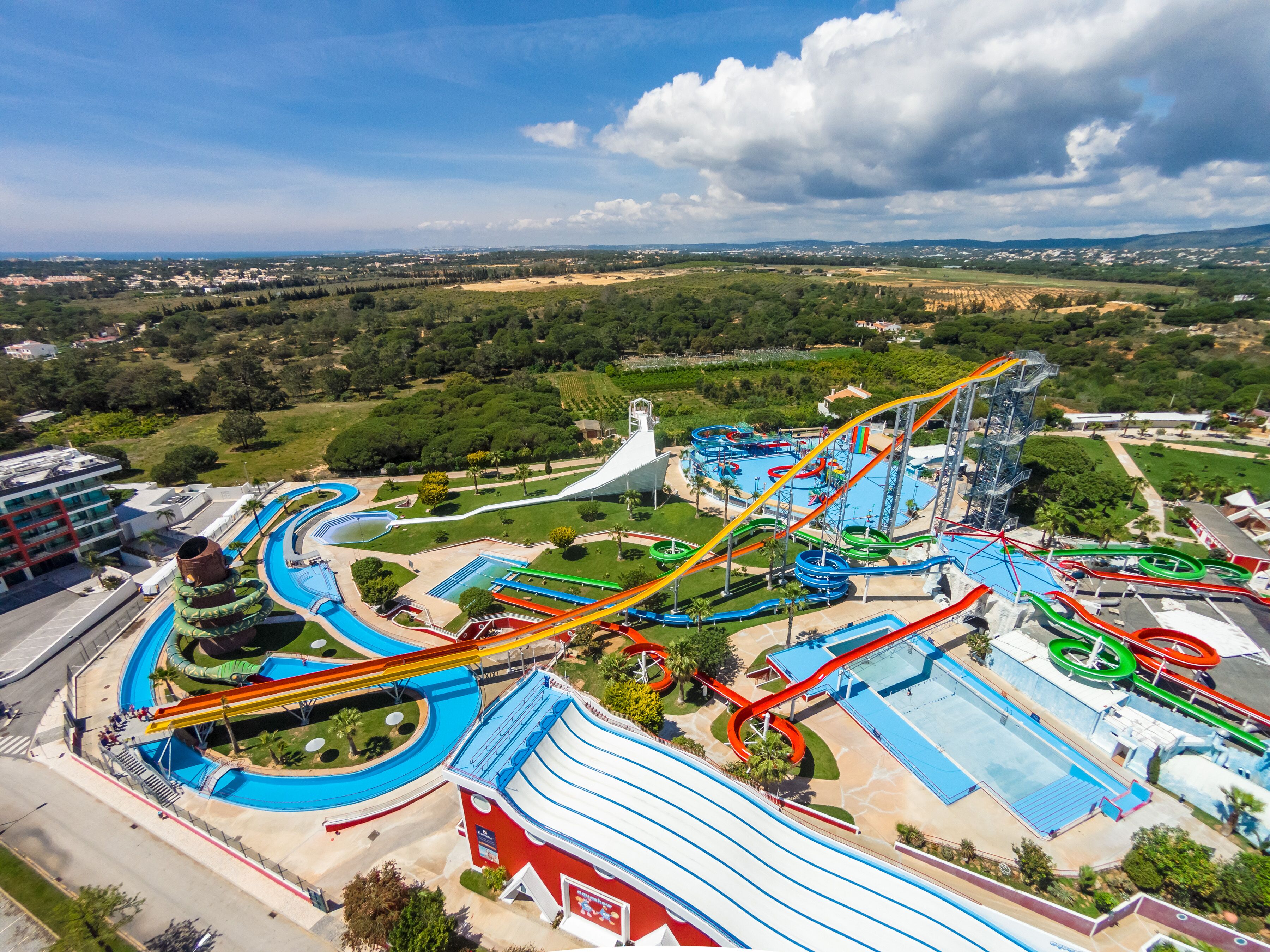 A water park with multiple slides and pools, surrounded by greenery and a distant cityscape.