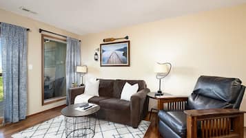 A living room with a brown sofa, a black leather chair, a coffee table, and a lamp. There is a window with curtains and a framed picture on the wall.