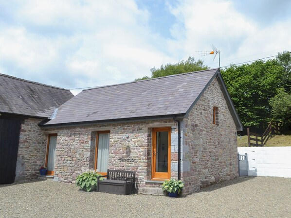A stone building with a sloped roof, a wooden door, and a small porch with benches and potted plants.