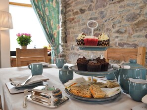 A three-tiered stand with cupcakes and sandwiches, a teapot, cups, and a small cake on a table with a stone wall background.