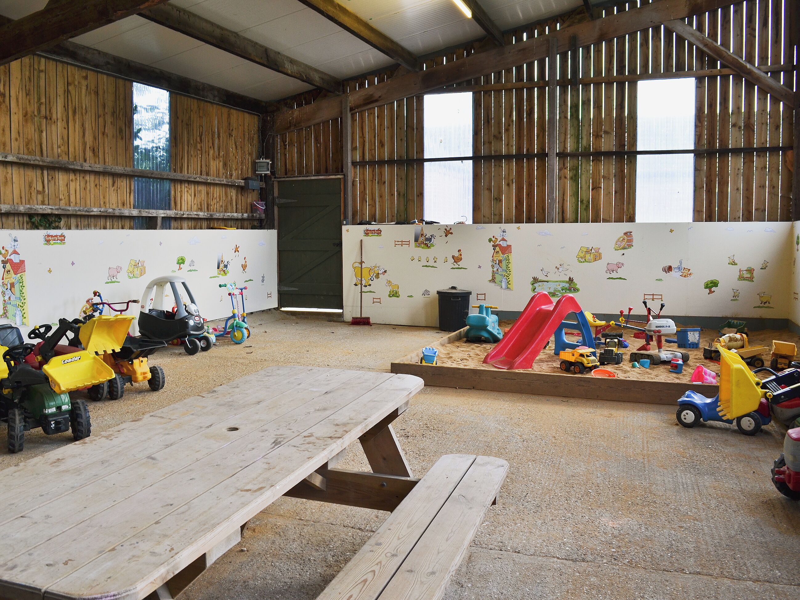 A spacious indoor play area with wooden walls, a large table, and various children's toys.