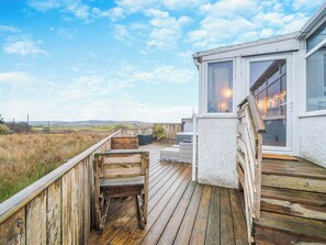 A wooden deck with outdoor seating, a white building with large windows, and a view of the countryside.