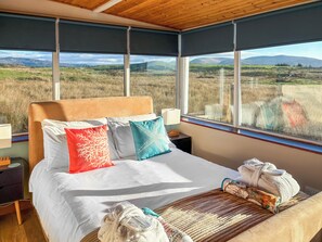 Une chambre avec un grand lit, de nombreux oreillers, une table de chevet avec une lampe, et une vue sur un paysage verdoyant par de grandes fenêtres.