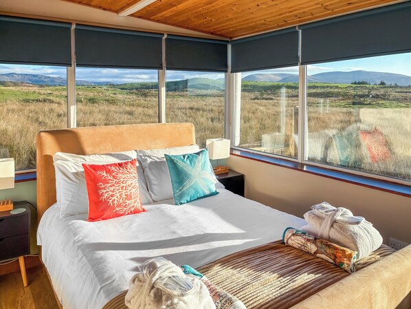 A bedroom with a large bed, multiple pillows, a bedside table with a lamp, and a view of a grassy landscape through large windows.