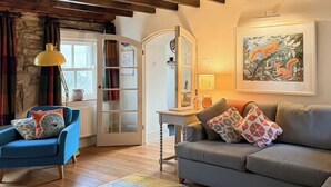 Ein Wohnzimmer mit blauem Sessel, grauem Sofa, Teppich mit Muster, Holzboden, weißer Tür mit Glasfeldern und einem Gemälde an der Wand.