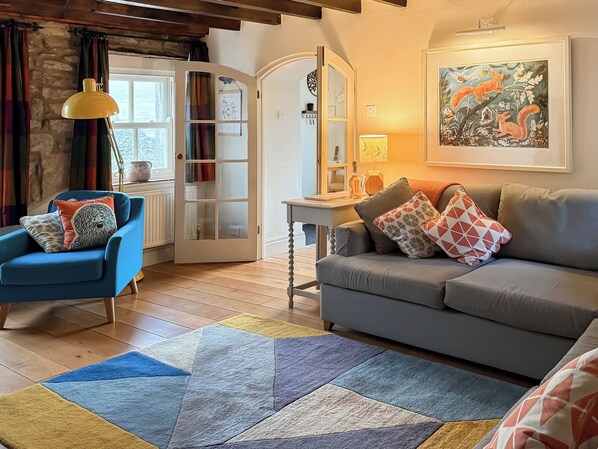 A living room with a blue armchair, a grey sofa, a patterned rug, a wooden floor, a white door with glass panels, and a painting on the wall.