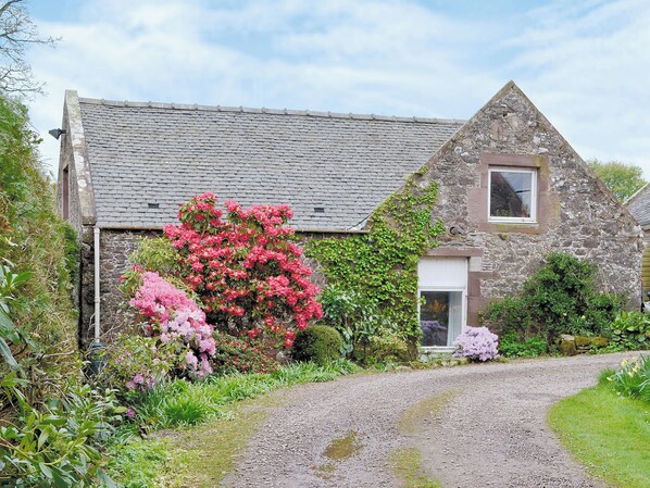 Una casa de piedra con entrada de grava, rodeada de exuberante vegetación y plantas con flores.