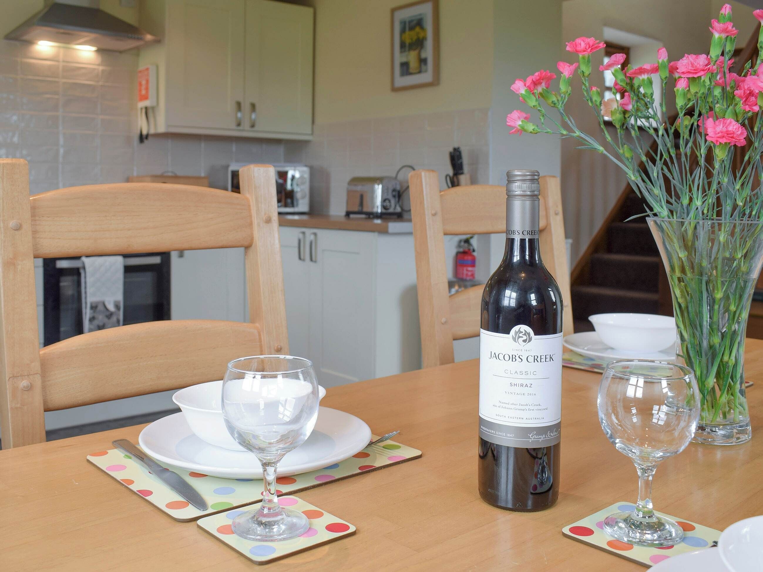 Une table à manger avec une bouteille de vin Jacob's Creek, deux verres à vin et un vase rempli de fleurs roses.