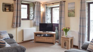 Een woonkamer met een televisie op een houten standaard, een kleine tafel met een bloemenvaas en een bank.