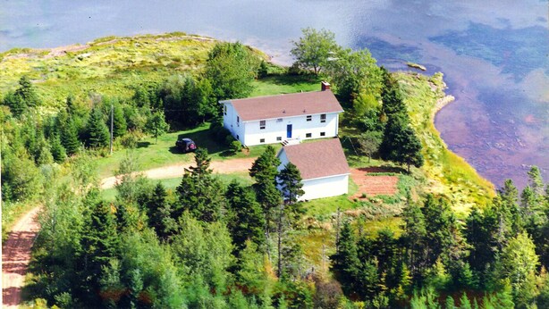 Une maison blanche à deux étages, surmontée d’un toit rouge, entourée d’une forêt dense et bordée d’un plan d’eau.