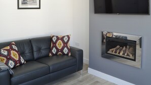 A modern living room with a dark leather sofa, two patterned throw pillows, a framed black and white photo on the wall, a flat-screen TV mounted above a fireplace with a log fire. - 3 bedroom accommodation in Amble (Amble)