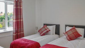 A room with two beds, each with a red blanket and a plaid pillow. There is a window with a red and white curtain, and a lamp on the nightstand. - 3 bedroom accommodation in Amble (Amble)