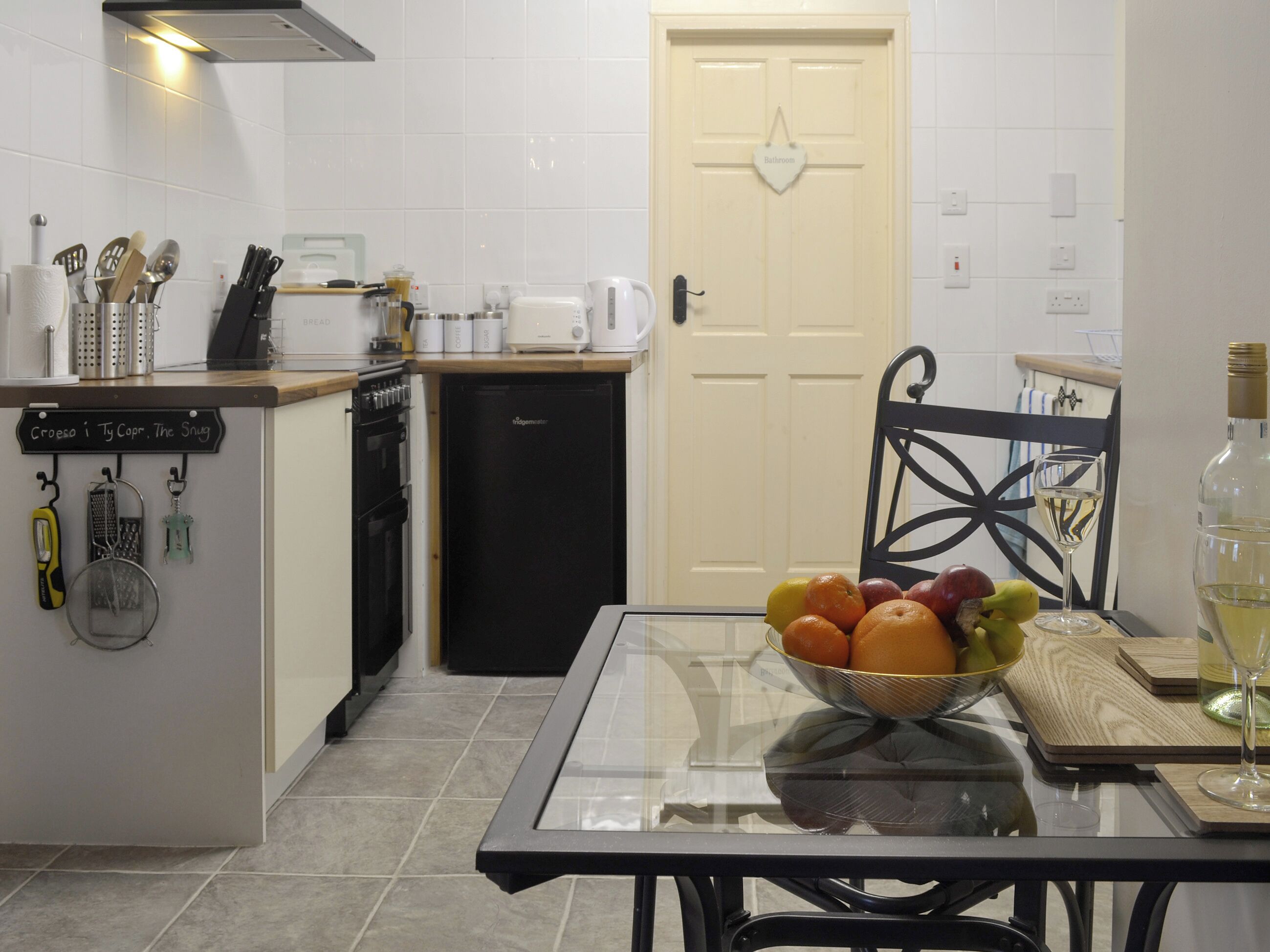 Una cocina con una mesa de vidrio, un cuenco de frutas, una copa de vino y una botella de vino.