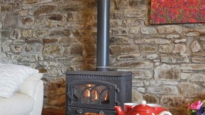 Une pièce chaleureuse avec un mur en pierre, un poêle à bois noir, un canapé blanc et une table sur laquelle se trouvent une théière, des tasses et une miche de pain.